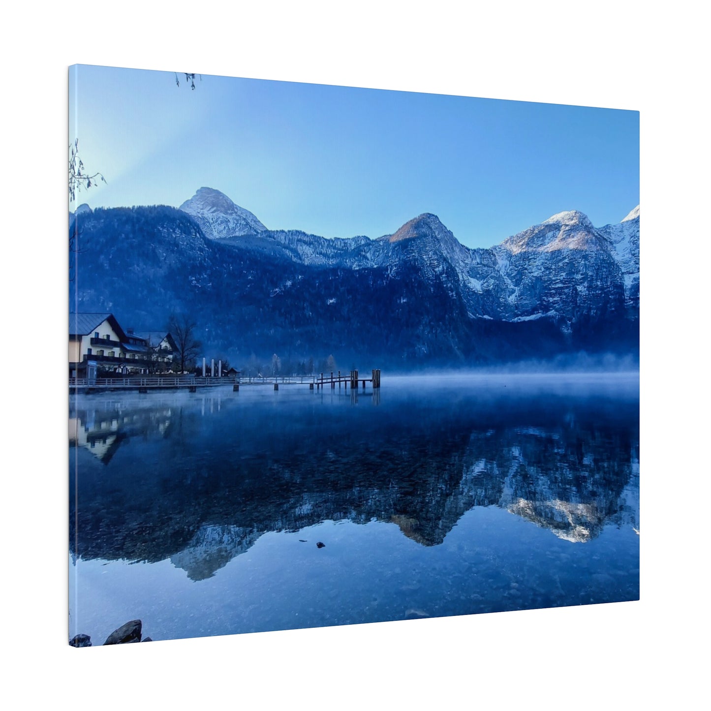 Calm lake waters reflecting snowy mountains and a village on the shore, under a bright blue sky. The tranquility of the scene is enhanced by the clear reflections.