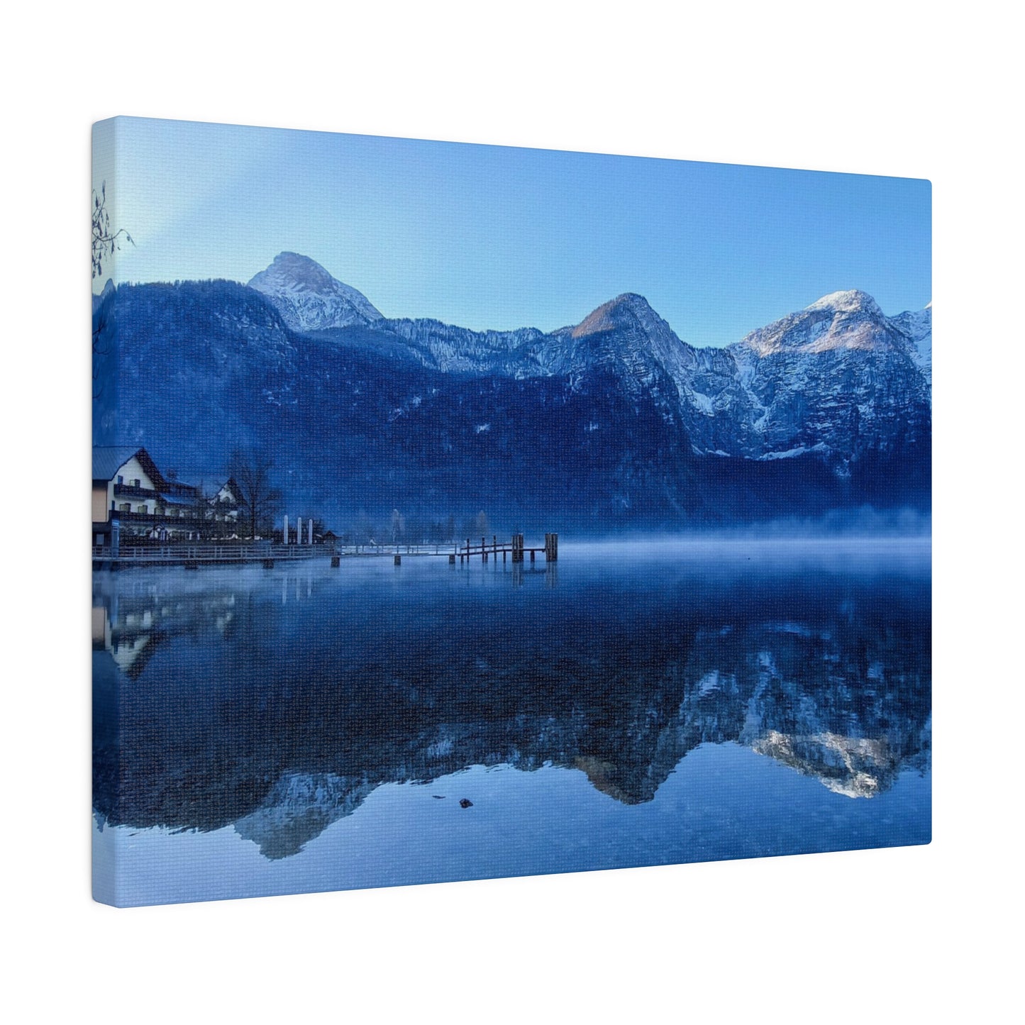 A calm winter lake with still waters mirroring snowy mountains and a village on the shore, under a bright blue sky. The reflection creates a peaceful view.