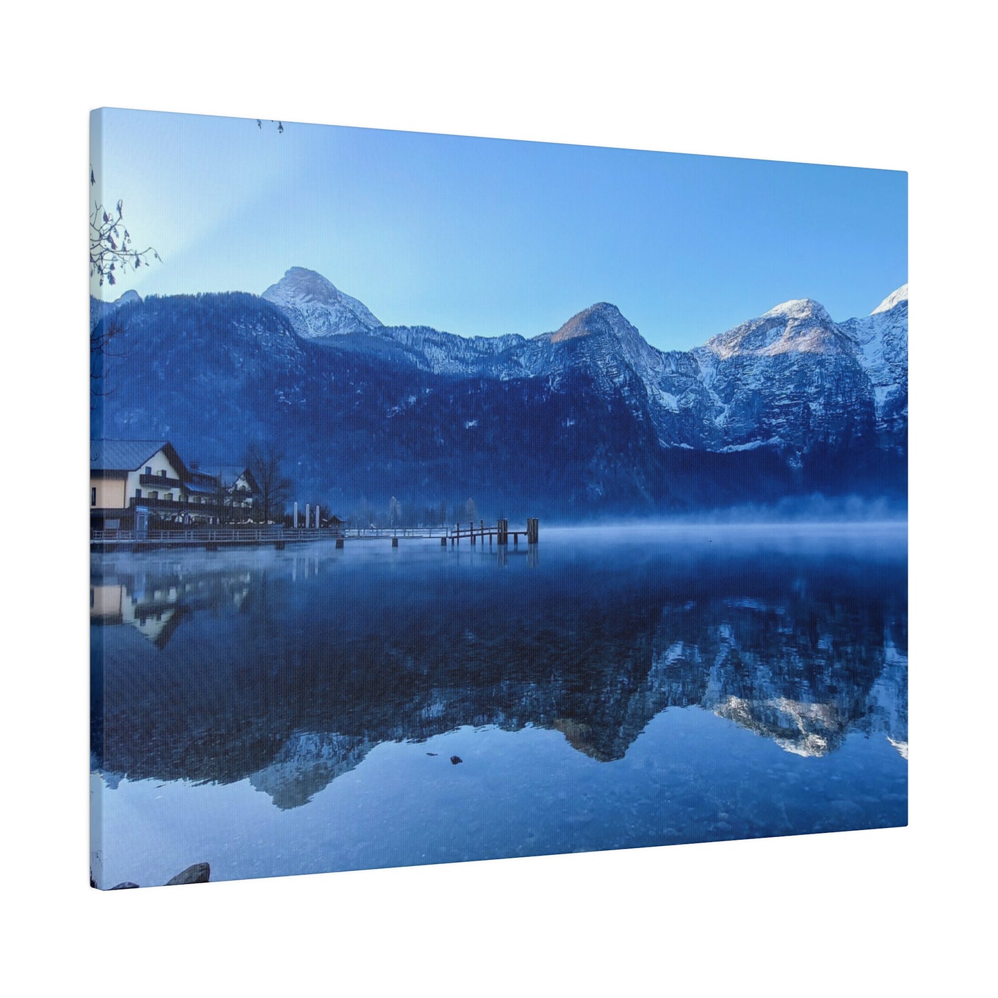 Clear calm lake waters reflecting snowy mountains and a village on the shore, under a bright blue sky. The reflection creates a peaceful winter scene.