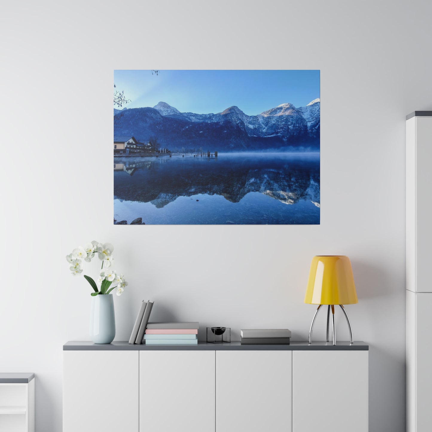 Clear lake waters reflecting snowy mountains, with a village on the left and a bright blue sky. The stillness of the water enhances the peacefulness of the scene.