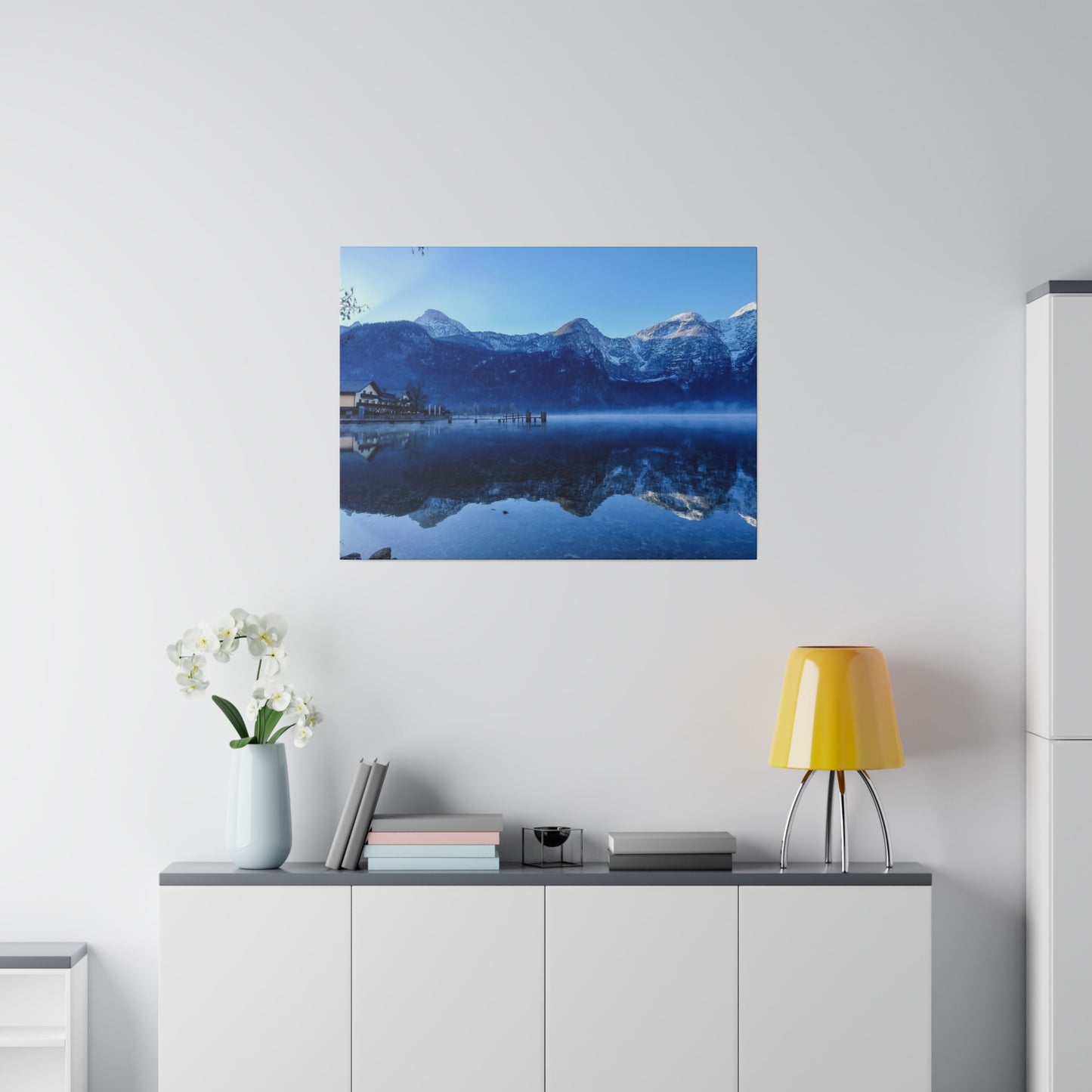 A mountain lake with calm waters reflecting snowy peaks, a village on the shore, and a clear blue sky. The reflection in the water adds to the tranquility of the scene.