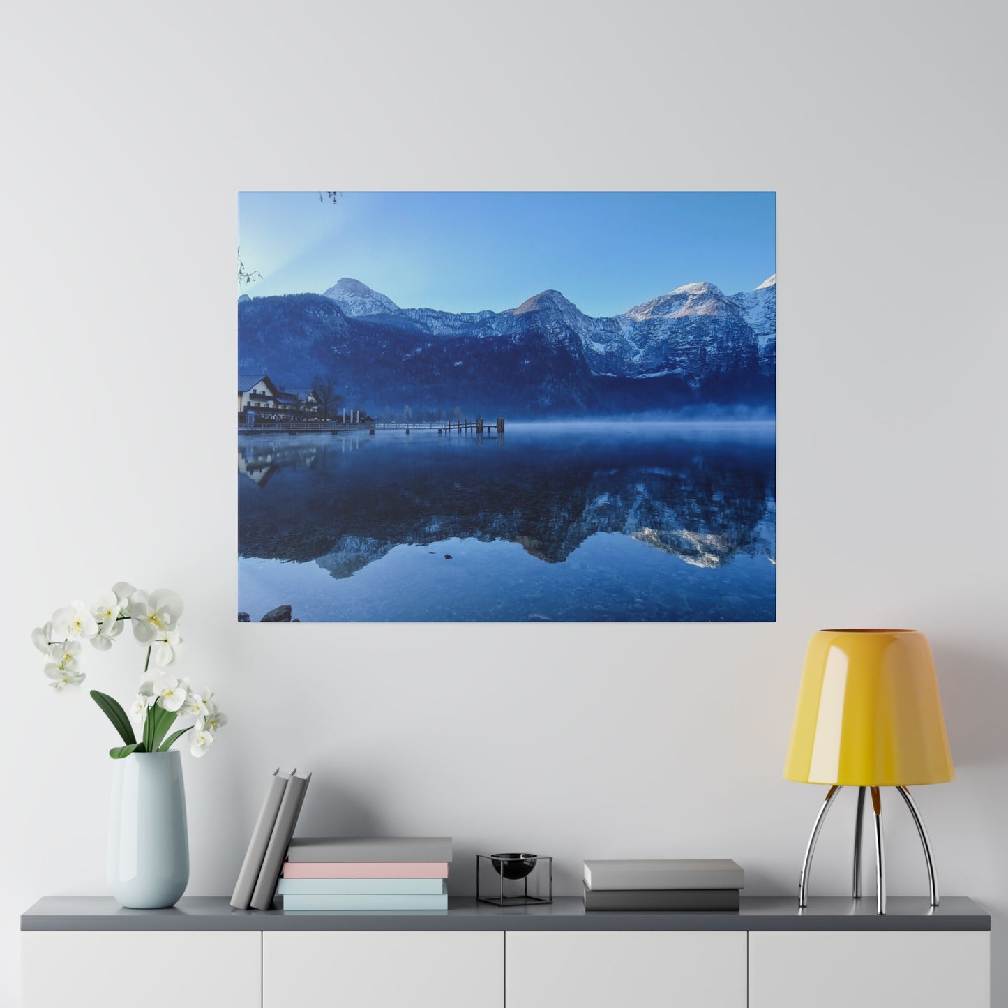 A peaceful lake view showing calm waters reflecting snowy peaks and a village with a dock on the shore. The scene is set against a backdrop of a bright blue sky.