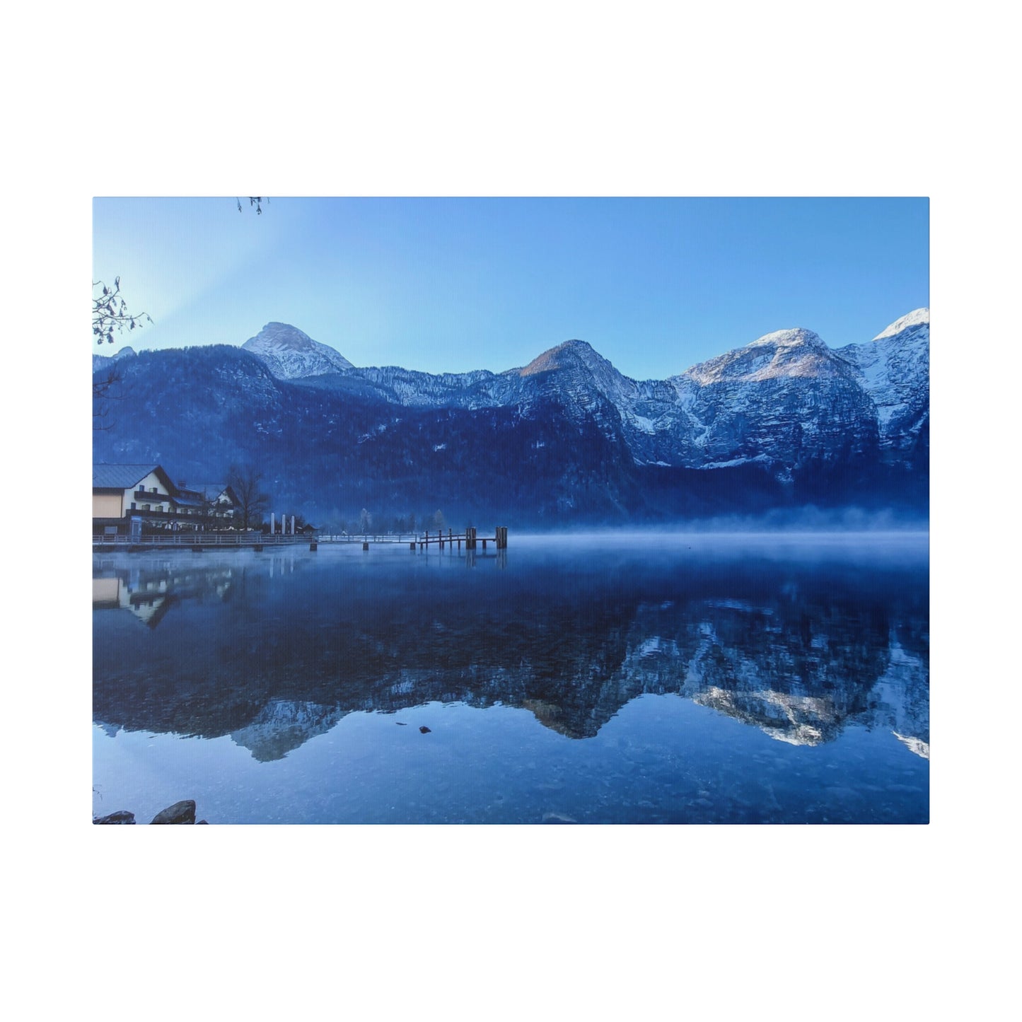 A peaceful winter lake view with calm waters reflecting snowy mountains, a village on the shore, and a bright blue sky. The scene is serene and beautiful.