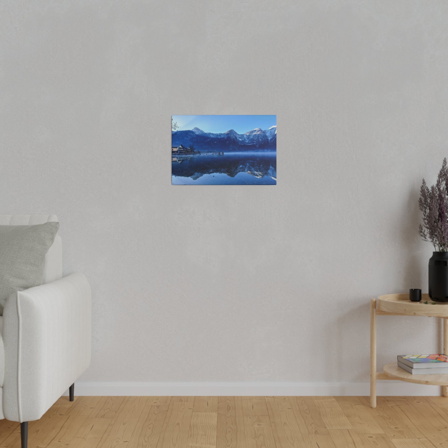 A peaceful winter lake with calm waters reflecting snowy mountains, a village on the shore, and a bright blue sky. The stillness of the water creates a perfect mirror image.