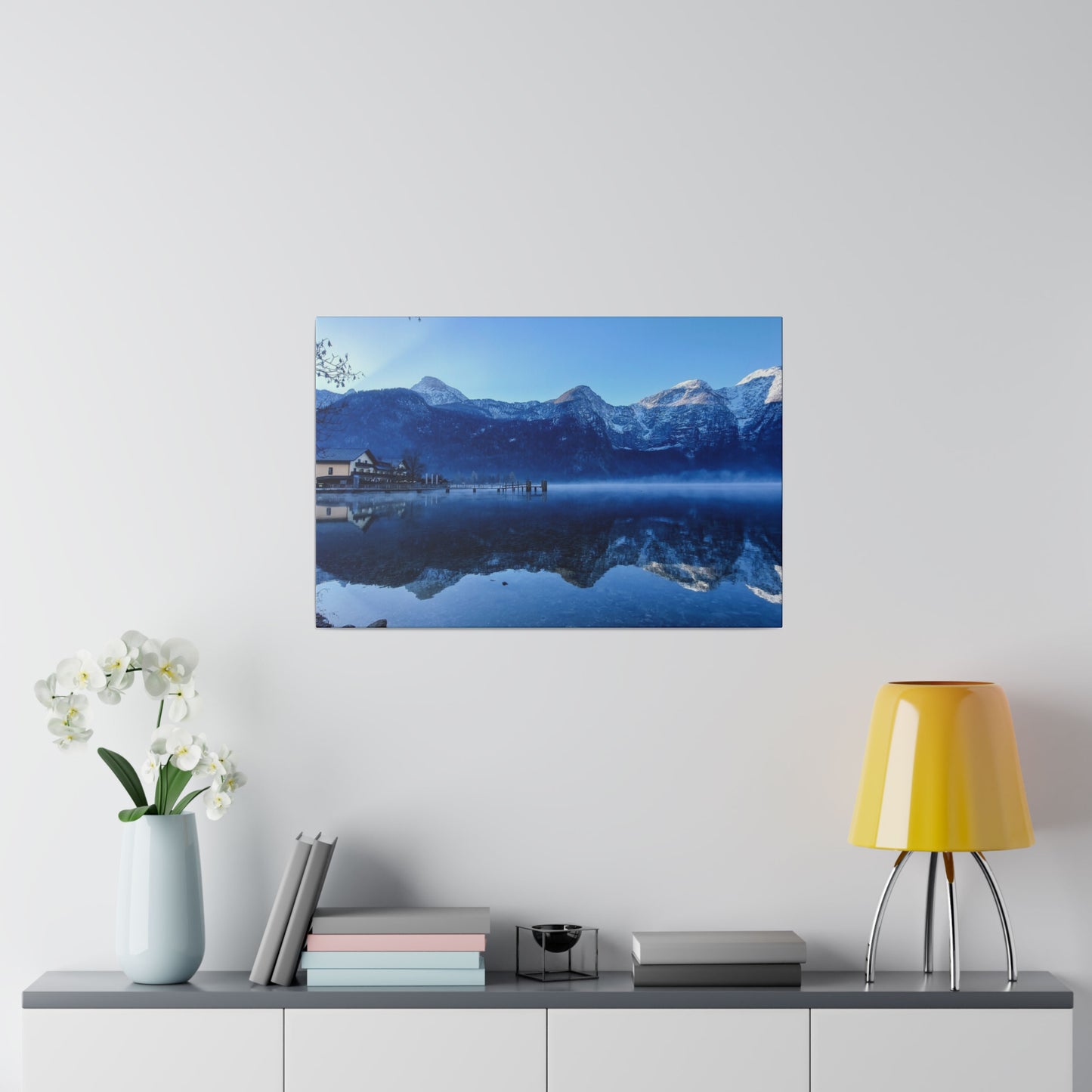 Reflective lake with snowy peaks and a village on the left, under a clear blue sky. The still waters create a perfect mirror image of the snow-covered mountains.