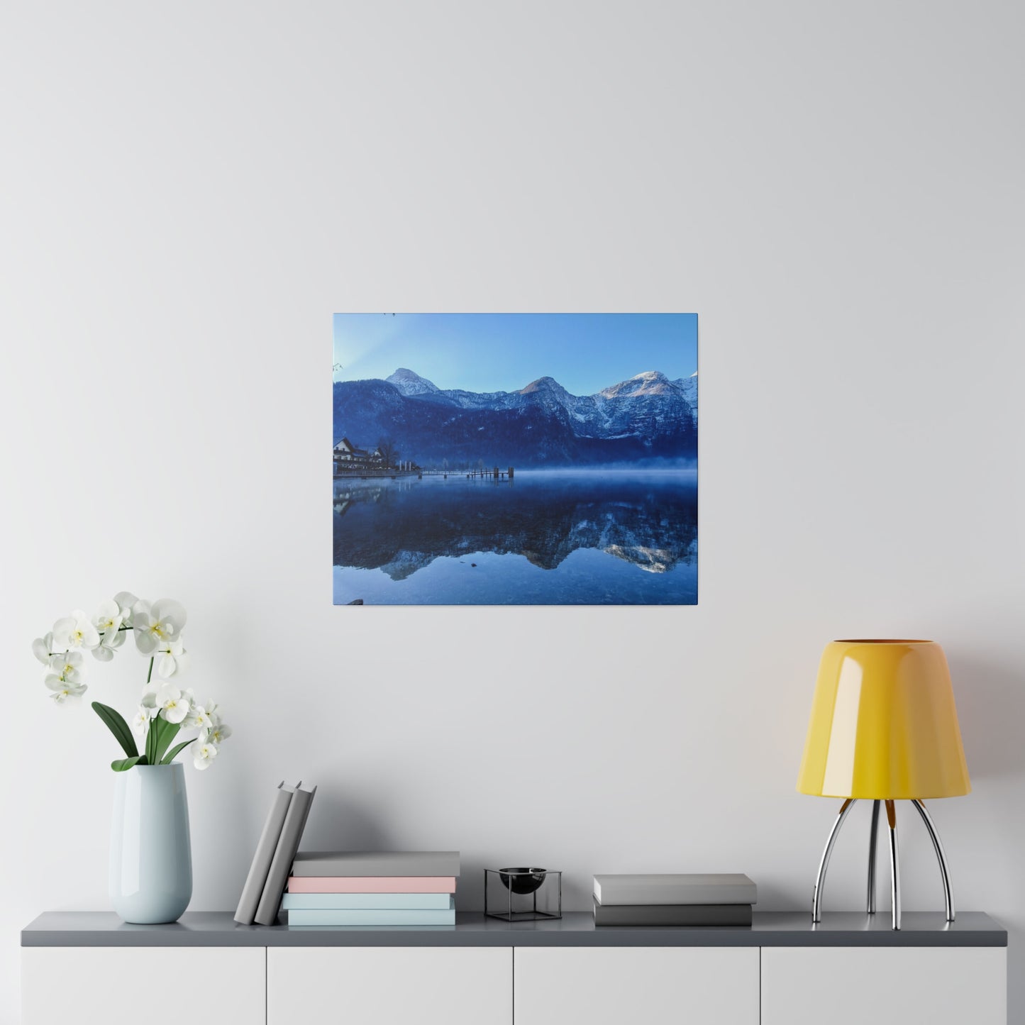 A scenic snowy lake with calm waters reflecting the mountains, a village on the shore, and a clear blue sky. The reflection adds to the picturesque beauty.