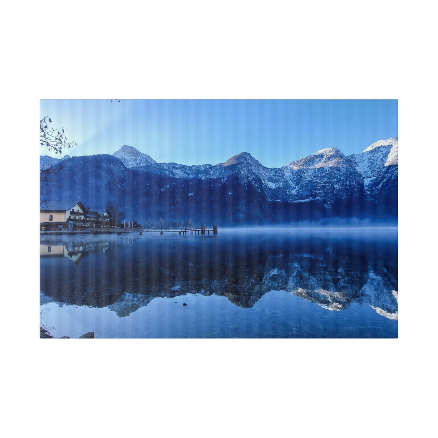 A serene lake in winter with calm waters reflecting snowy mountains, a village on the shore, and a bright blue sky. The reflection creates a tranquil scene.