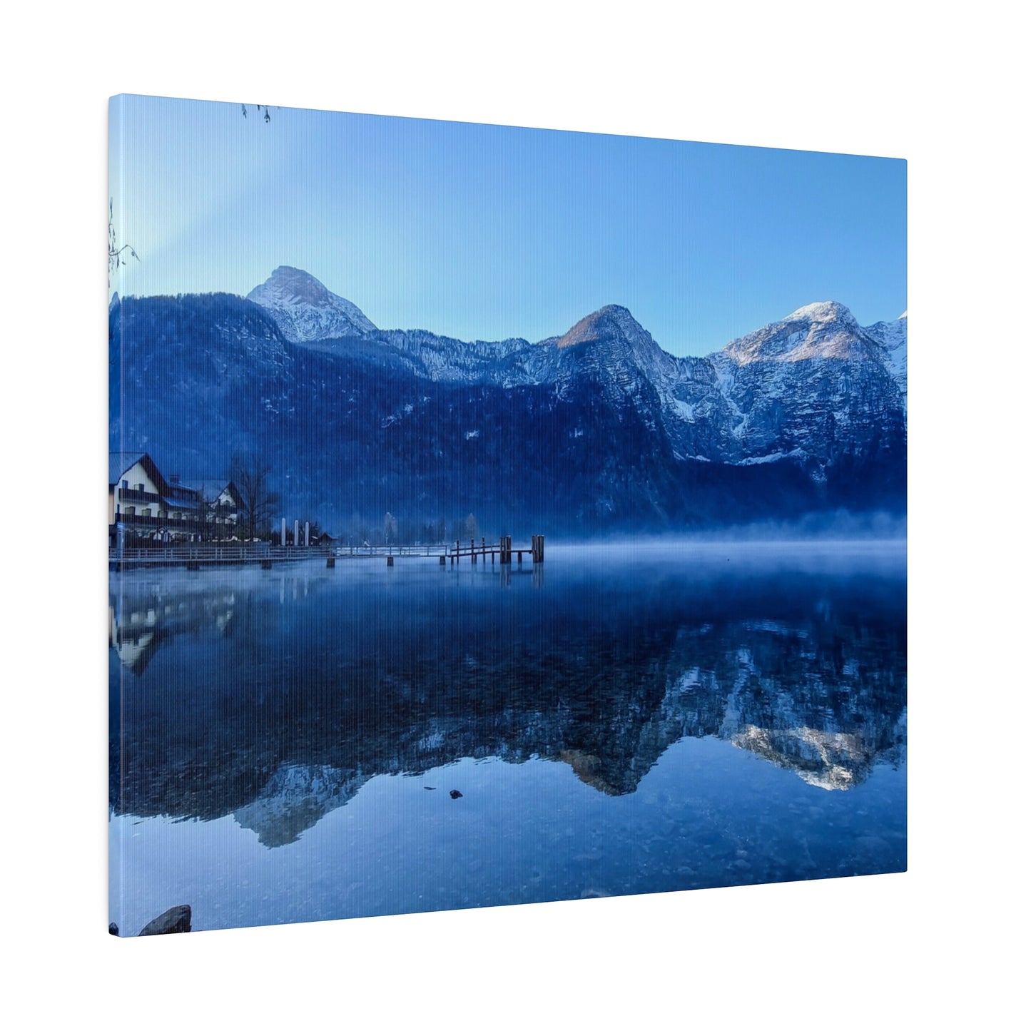 A serene mountain lake with calm waters reflecting snowy peaks, a village on the left, and a clear blue sky. The reflection creates a mirror image, enhancing the tranquility.