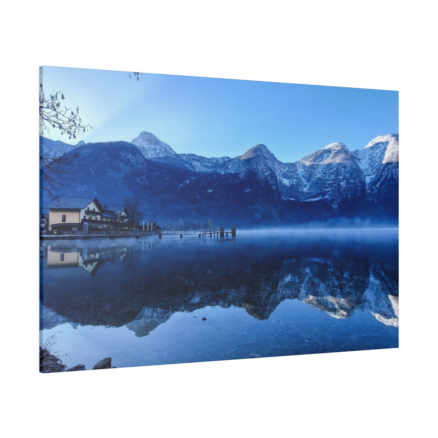 A serene winter reflection with calm lake waters showing snowy mountains, a village on the shore, and a bright blue sky. The still water creates a peaceful image.