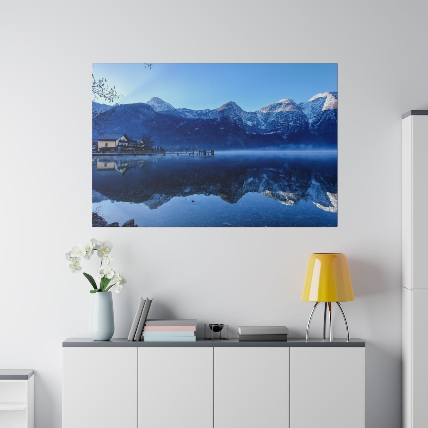 Snowy mountains reflected in a calm lake, with a village on the shore and a clear blue sky. The serene waters add to the tranquil winter scene.