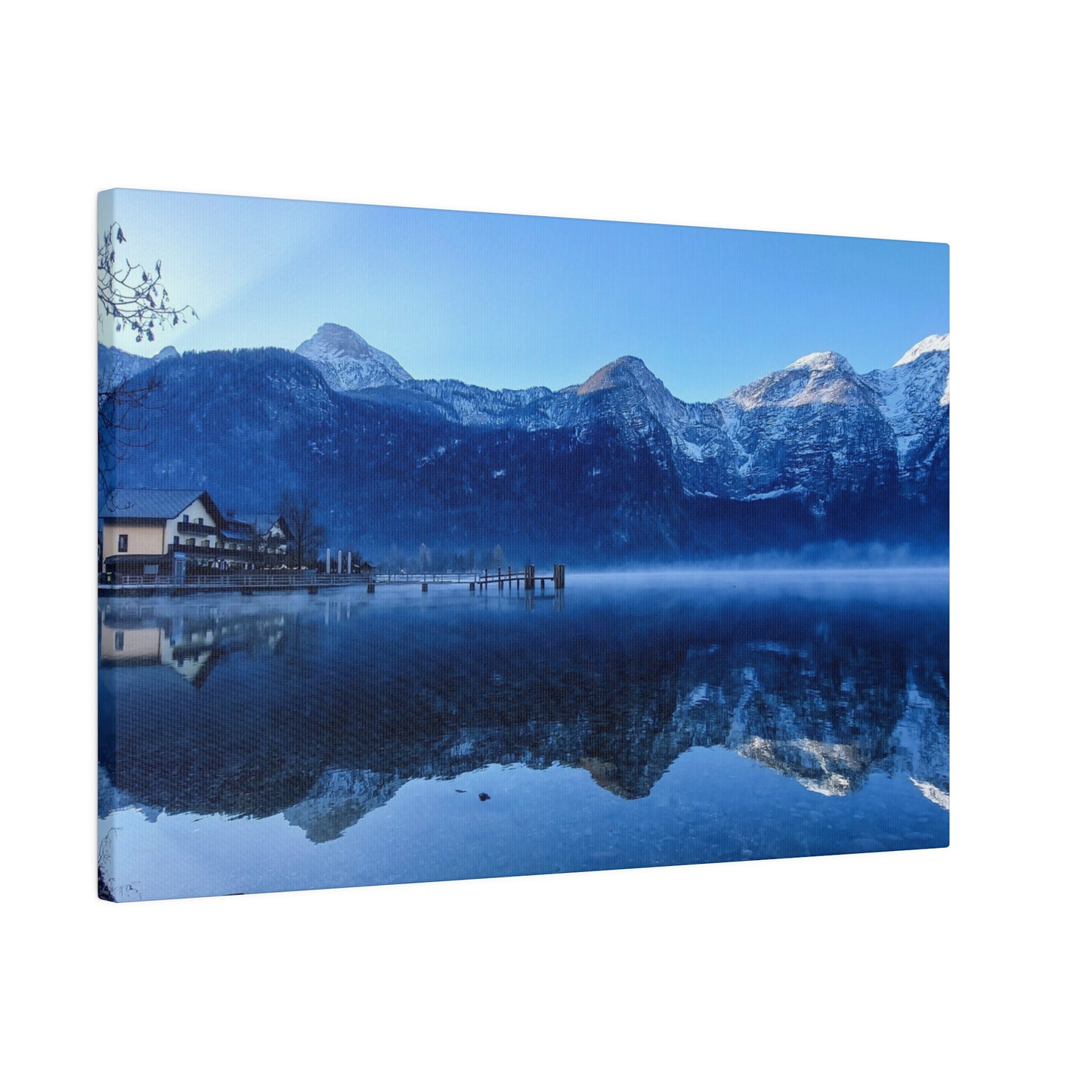 A snowy lake view with calm waters mirroring the mountains, a village on the shore, and a clear blue sky. The reflection in the lake creates a serene winter landscape.