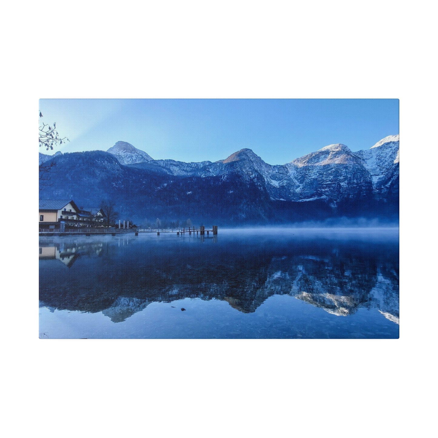 A snowy mountain lake with calm waters reflecting the peaks, a village on the shore, and a bright blue sky. The reflection creates a serene and peaceful scene.