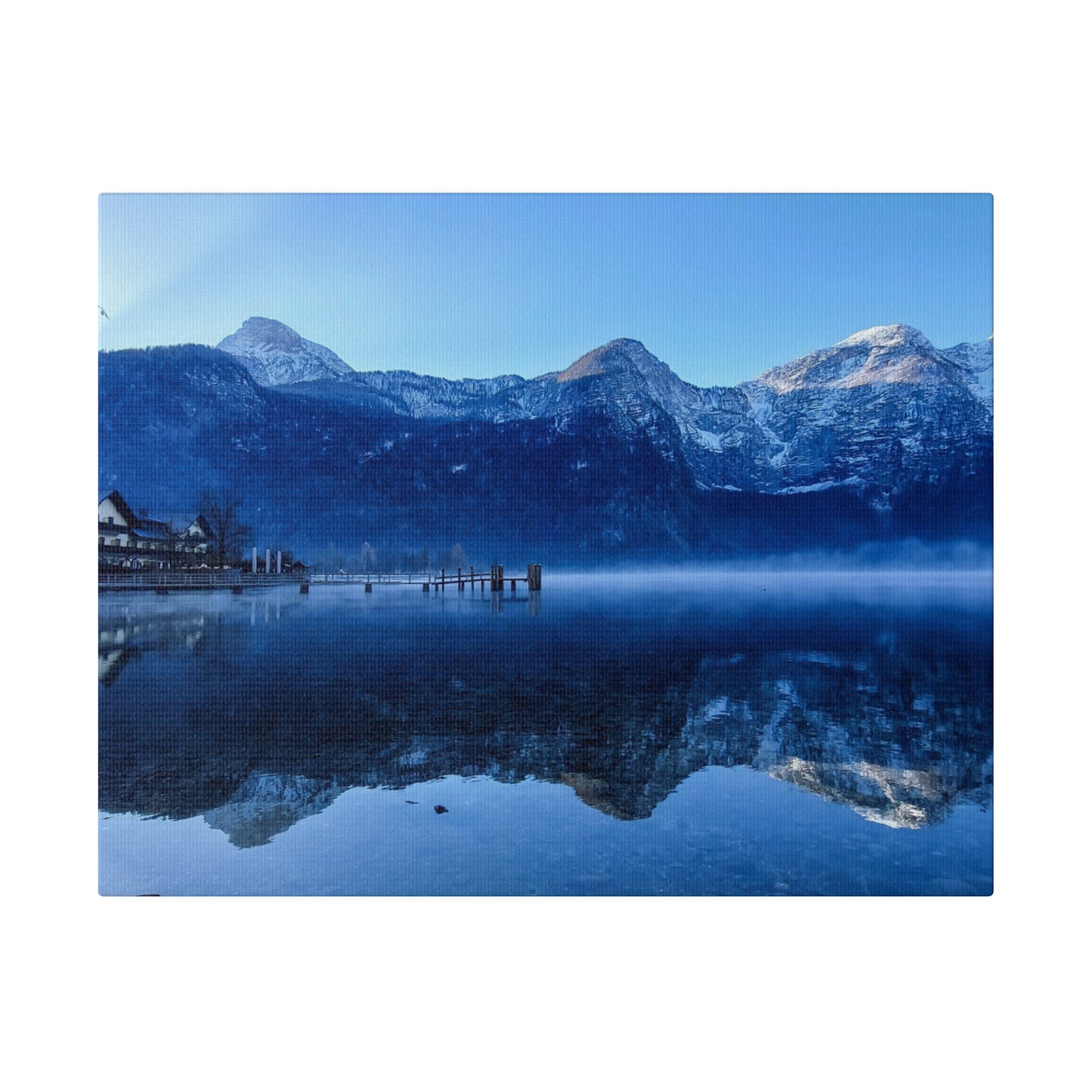 Snowy peaks reflected in a calm lake, with a village on the shore and a bright blue sky. The reflection creates a peaceful and serene winter landscape.
