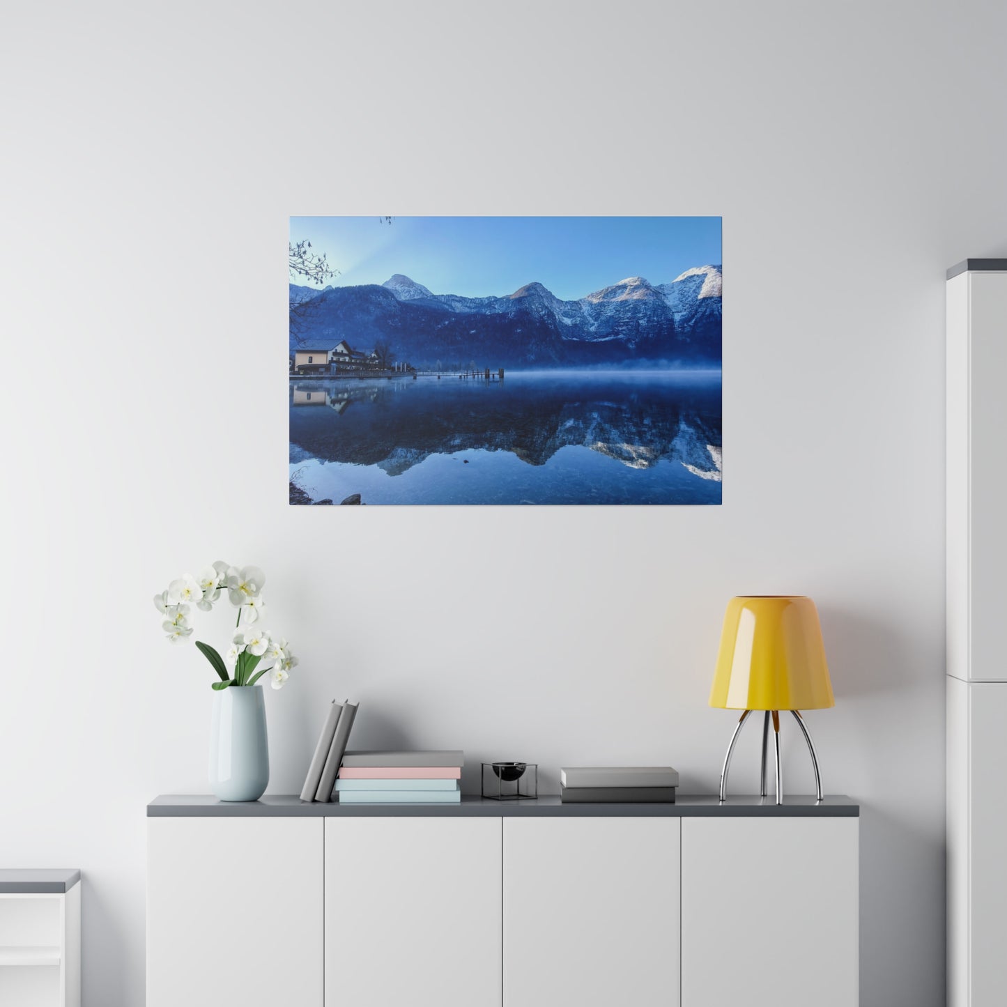 Snowy peaks reflected in a calm lake, with a village on the shore and a bright blue sky. The clear reflection in the water enhances the peacefulness of the landscape.