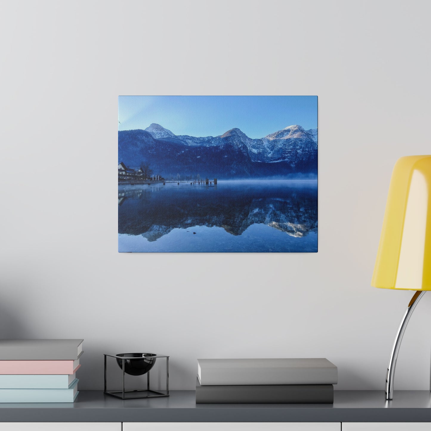 A still snowy lake with calm waters reflecting the mountains, a village on the shore, and a clear blue sky. The reflection adds to the tranquility.