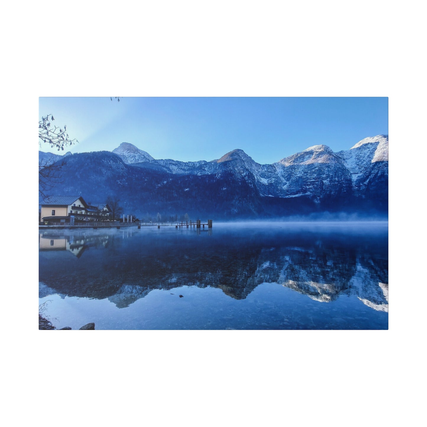 A tranquil reflective lake with calm waters showing snowy mountains, a village on the shore, and a bright blue sky. The reflection creates a serene winter scene.