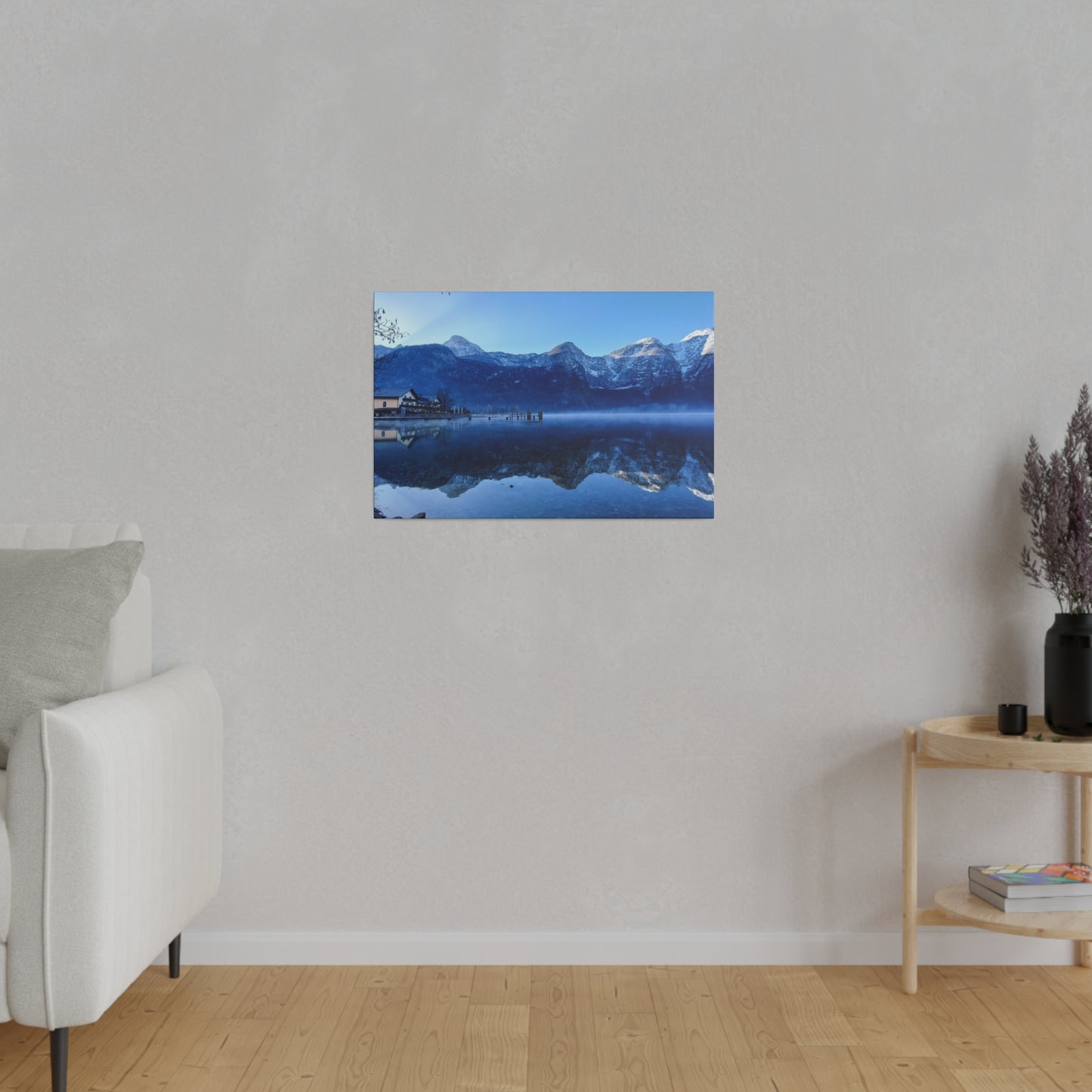 A winter lake landscape with still waters reflecting snowy mountains and a village, under a bright blue sky. The reflection in the lake adds to the peacefulness of the scene.