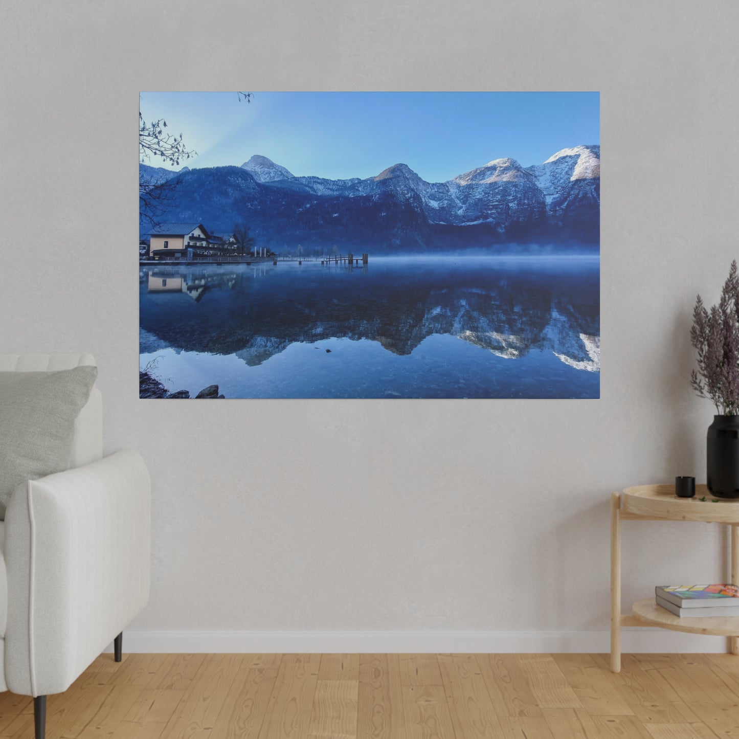 A winter reflective lake with still waters showing snowy mountains and a village on the shore, under a clear blue sky. The reflection adds to the calm atmosphere.