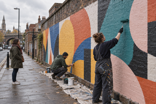 Two artists paint an abstract geometric mural on an urban brick wall while a passer-by watches from the pavement.