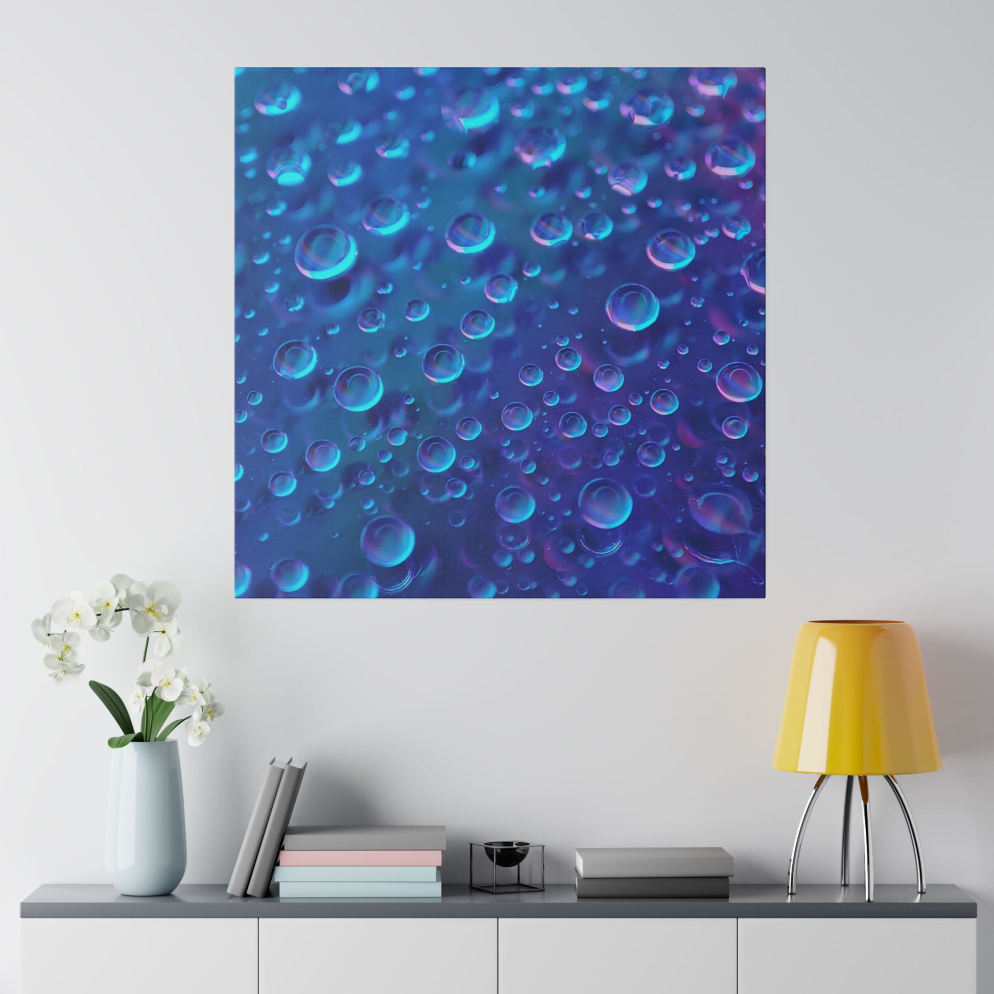 Blue abstract print of bubbles in varying sizes hangs on a white wall above a gray cabinet with a pastel vase, books, and a yellow table