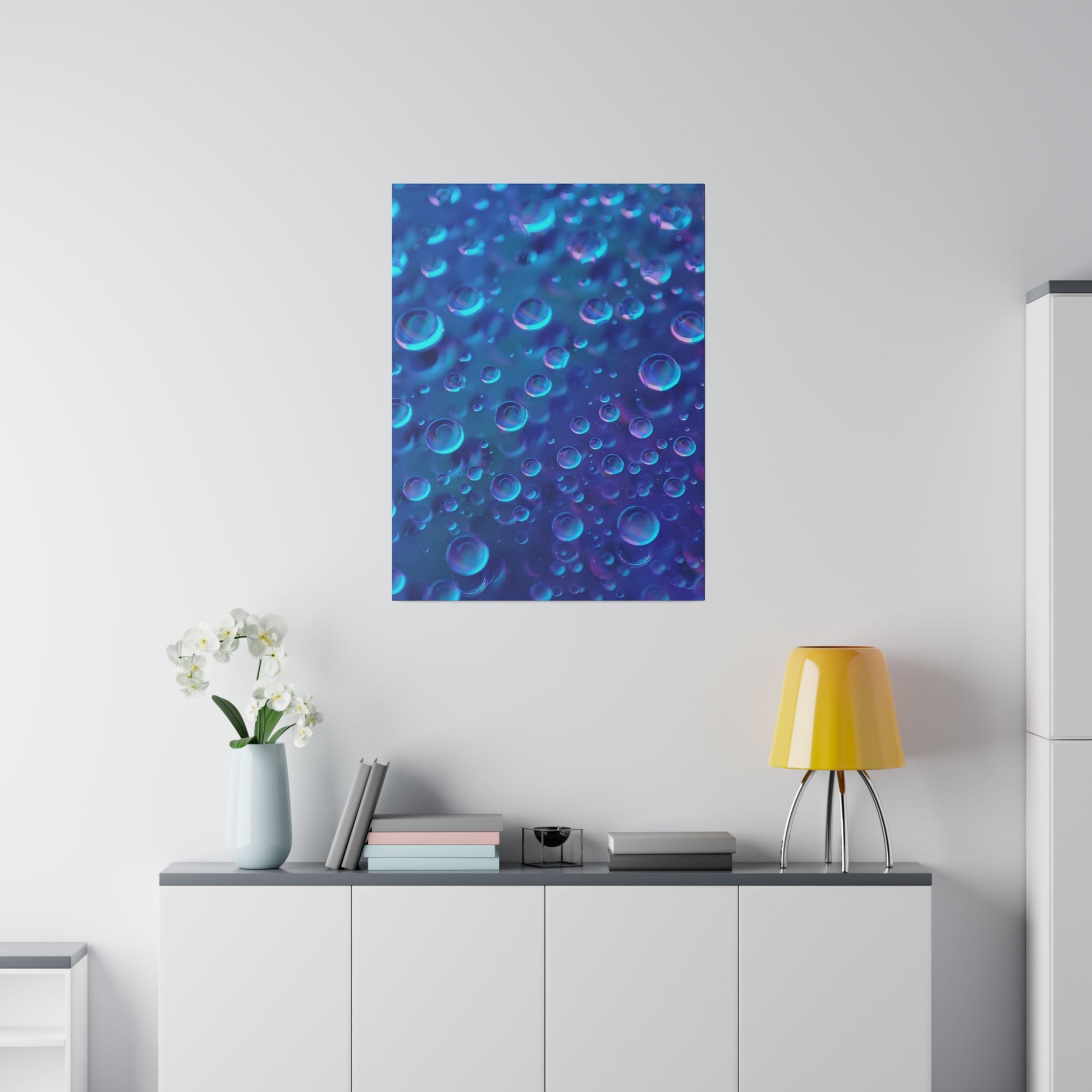 Blue poster with bubbles hangs on a white wall above a gray-topped white cabinet, beside a yellow lamp and a vase of white flowers.