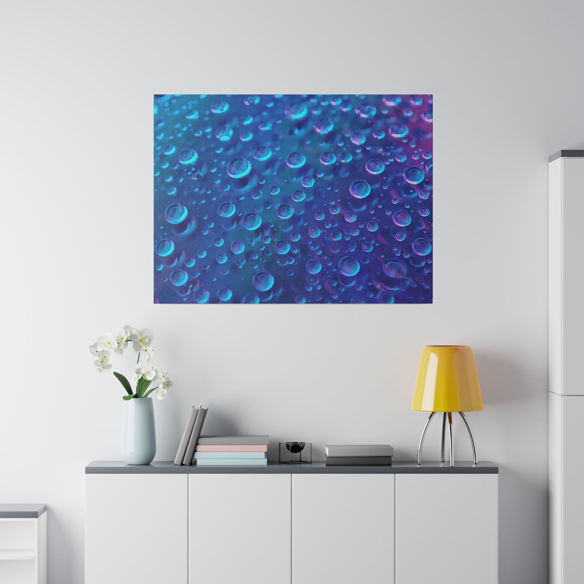 Blue and purple abstract bubble art hanging above a white cabinet with a vase of white flowers, books, and a yellow lamp.