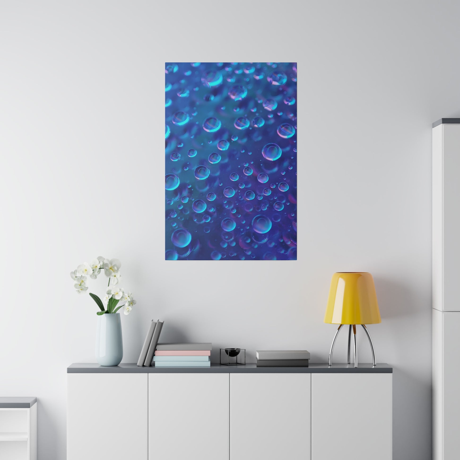 Blue poster of numerous bubbles hangs on a white wall above a white cabinet with a yellow lamp and books.