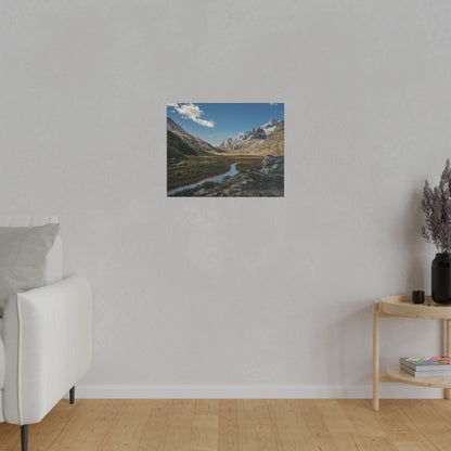 Small landscape poster of a mountain valley with a river, blue sky, and snow-capped peaks mounted on a light gray wall.