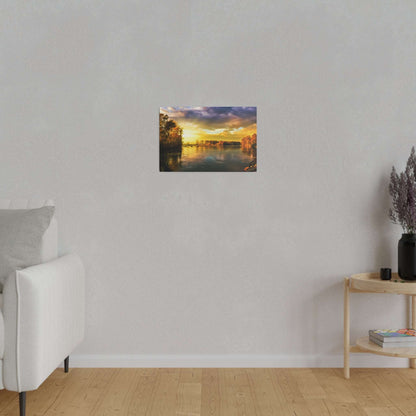 Small landscape painting of a sunset over a calm river with orange sky, mounted on a light gray wall above a wooden floor.