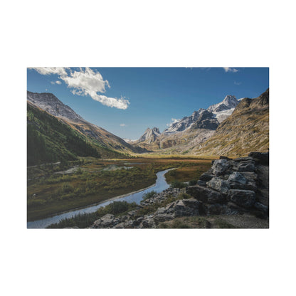 Alpine valley scene with a winding river through green meadows, rocky foreground, and snow‑capped mountains under a blue sky.