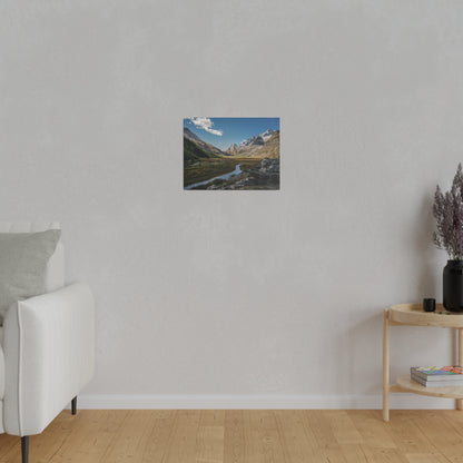 Small landscape wall art showing a mountain valley with a river, mounted on a light gray wall in a living room.
