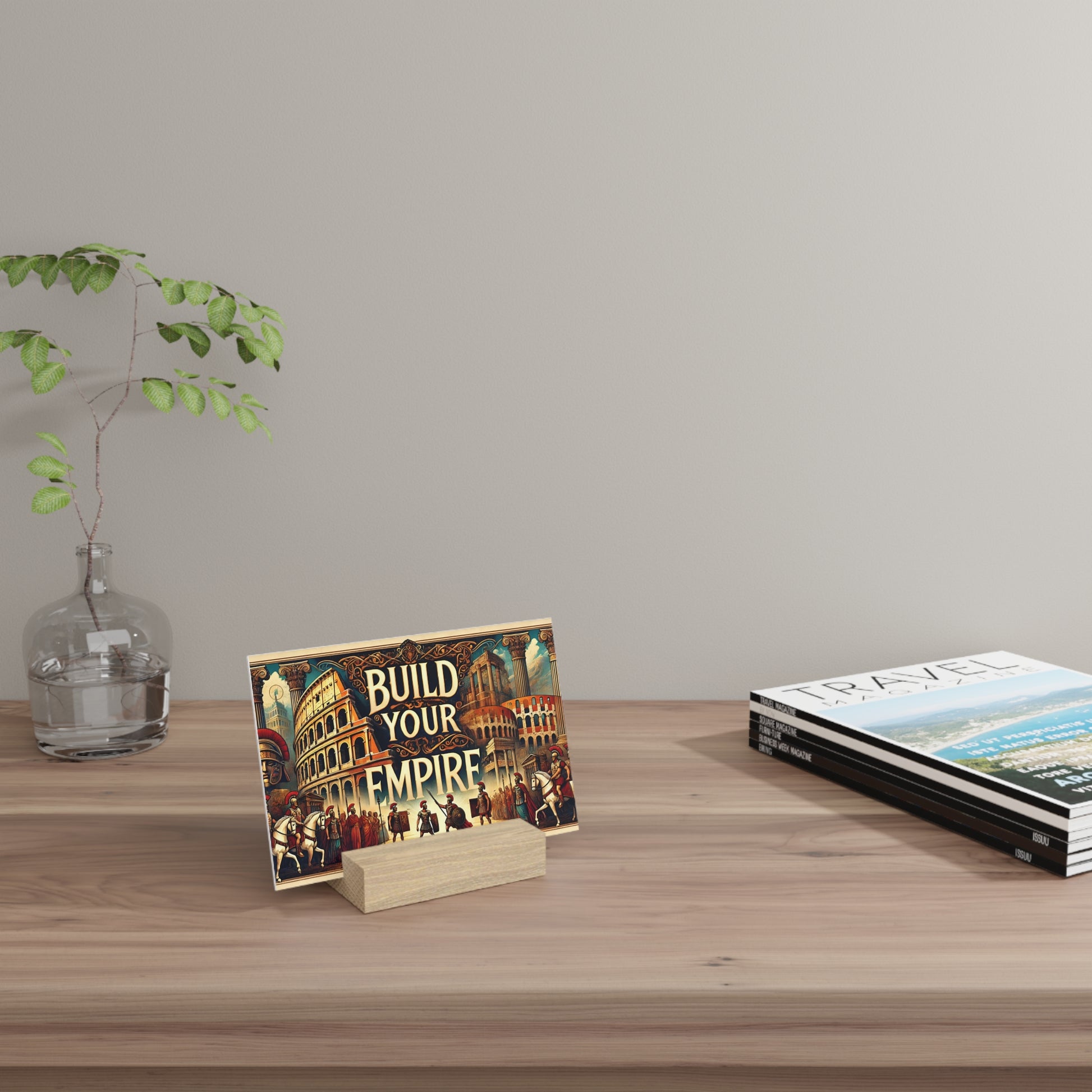 Colorful “Build Your Empire” poster on a wooden block stand sits on a light wood table beside a glass vase with green branches.
