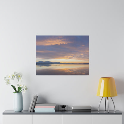 Square photo of a sunset over a calm lake with distant mountains, hung on a white wall.