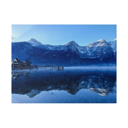 Snow-capped mountain range overlooking a calm lake with a wooden dock and small buildings on the left.