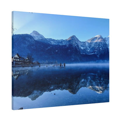 Blue-tinted landscape of a calm lake with snow-capped mountains, a small village on the left, and a wooden pier reflecting in the water.