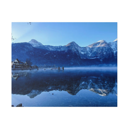 Snow-covered mountains rise behind a calm lake with a small wooden pier and a house-lined shore, mirror-reflected in the water.
