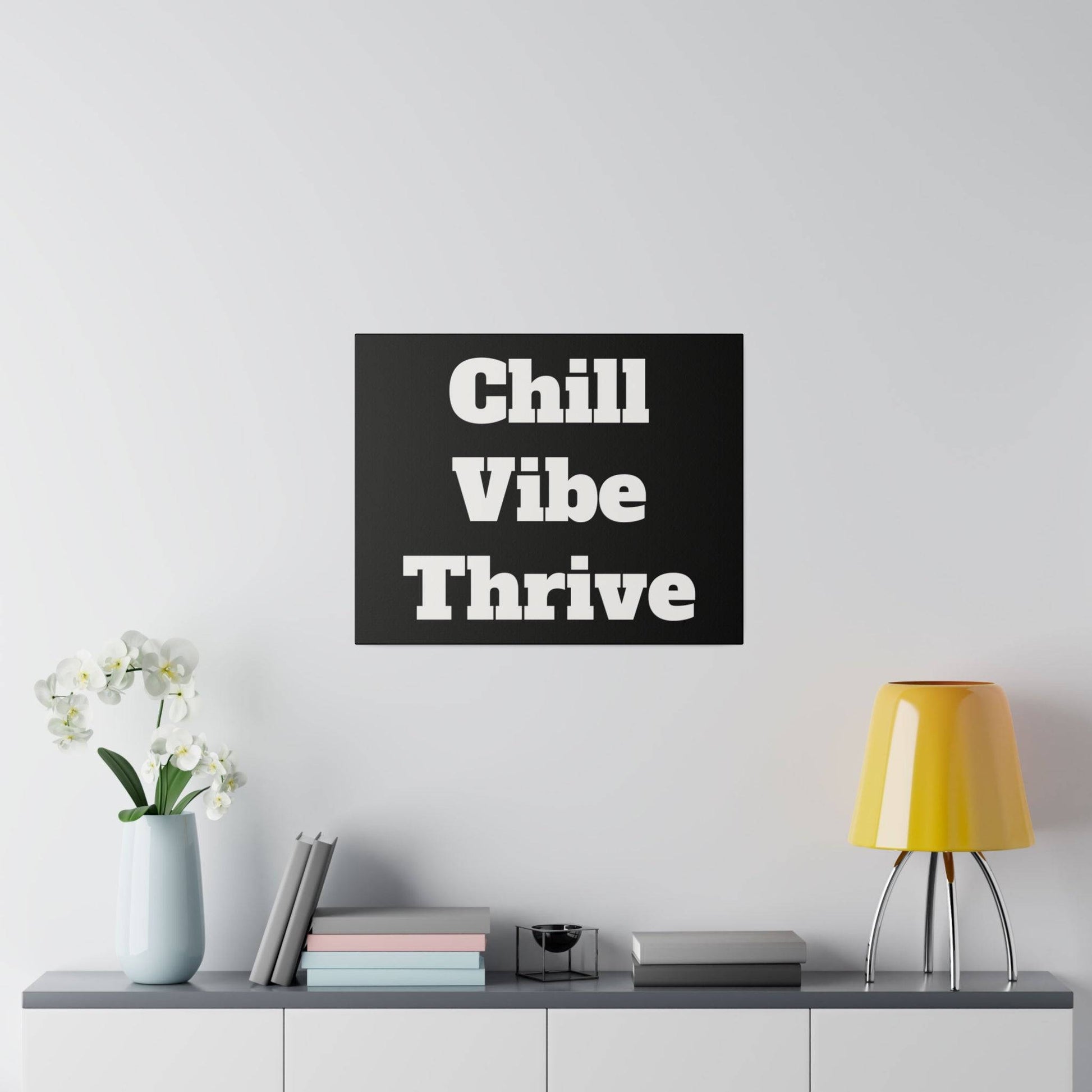 Black square wall art with large white text reading “Chill Vibe Thrive” hung above a white sideboard with a vase, books, and a yellow lamp.