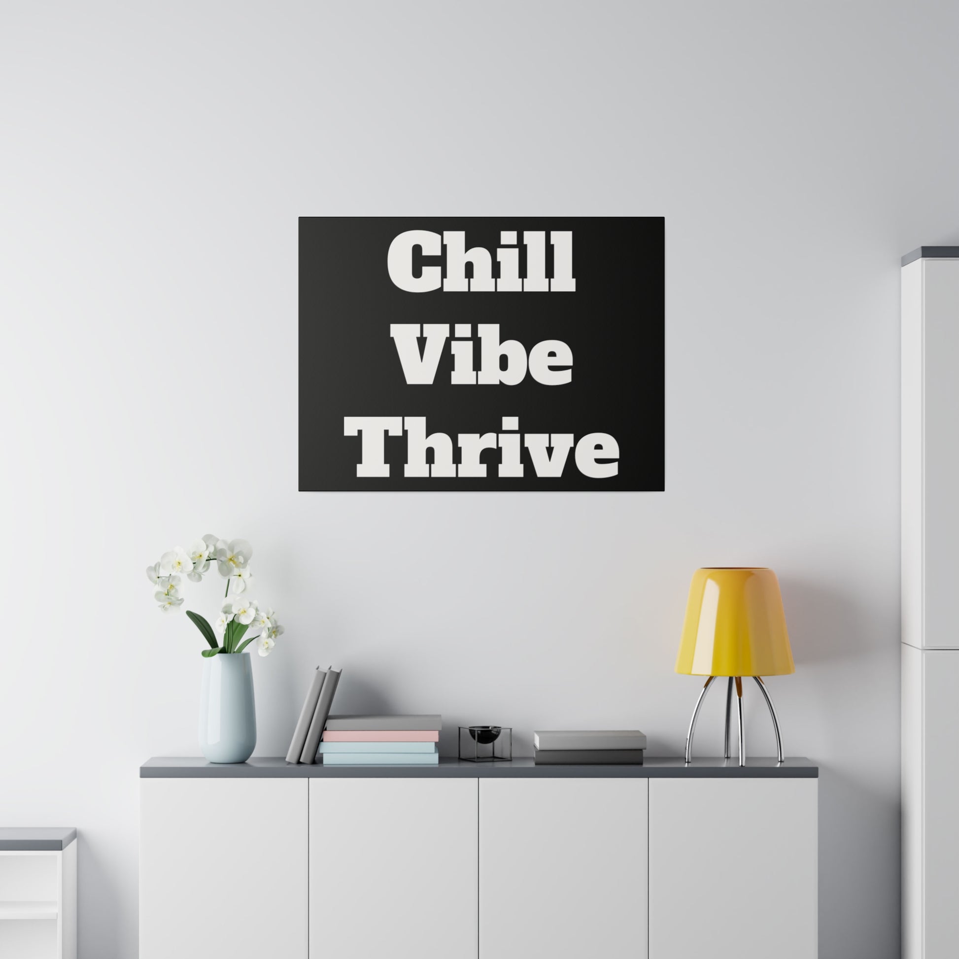Black wall art with bold white text “Chill Vibe Thrive” hangs above a white cabinet in a minimal living room with a yellow lamp and vase.