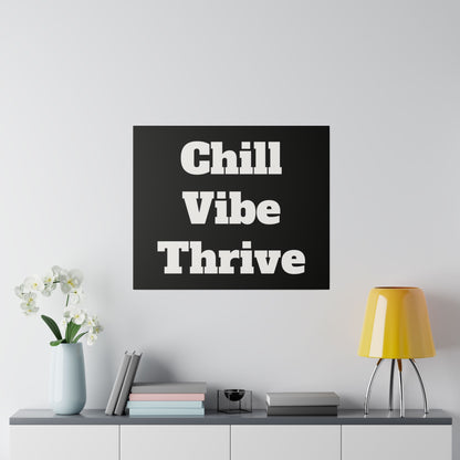 A black square poster with large white text reading "Chill Vibe Thrive" mounted on a light gray wall above a desk with a yellow lamp and