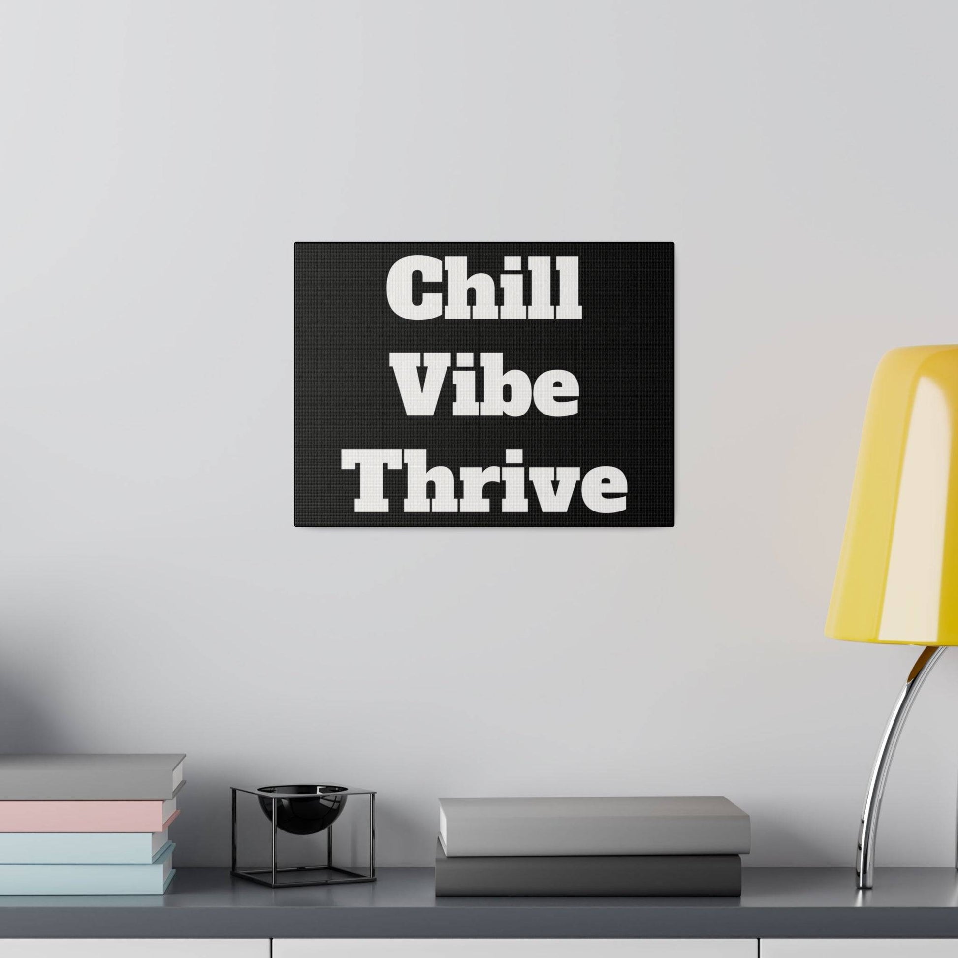 Black wall art with white text Chill Vibe Thrive hangs above a shelf with books, a small black bowl, and a yellow lamp.