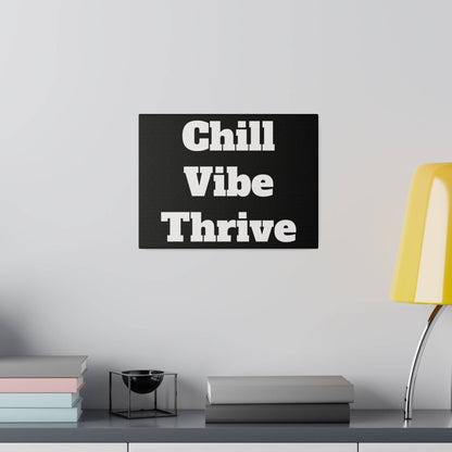 Black wall art with white text Chill Vibe Thrive hangs above a shelf with books, a small black bowl, and a yellow lamp.