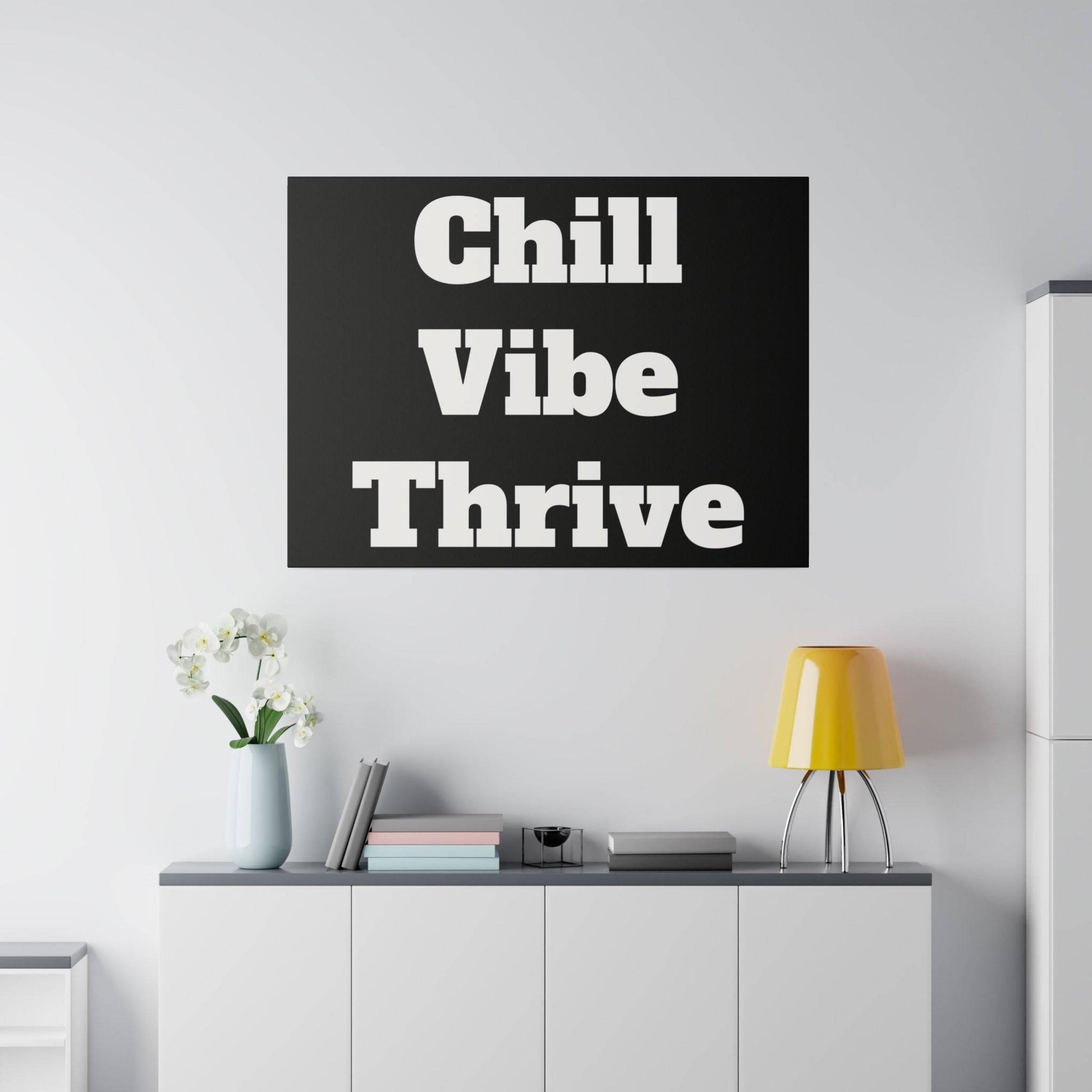 Black rectangular wall art with large white bold text reading “Chill Vibe Thrive” mounted above a white cabinet in a minimalist room.