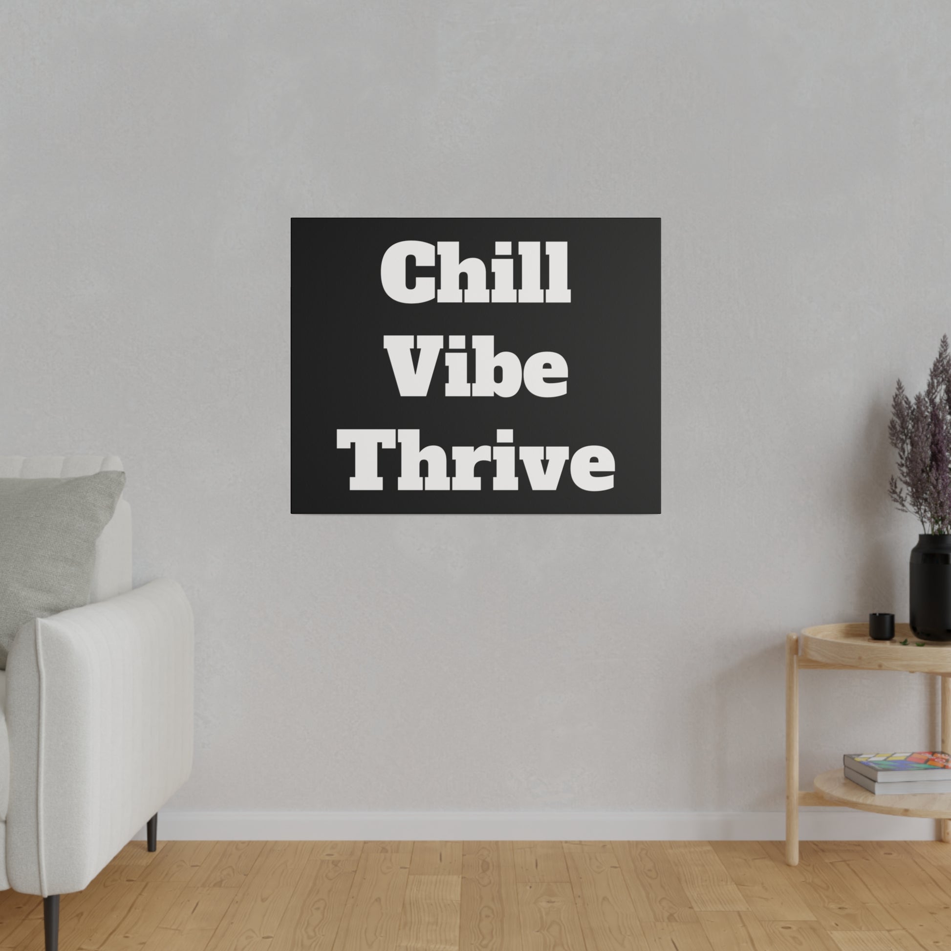Black square wall art with bold white text "Chill Vibe Thrive" hung on a light gray living room wall above a wooden floor.