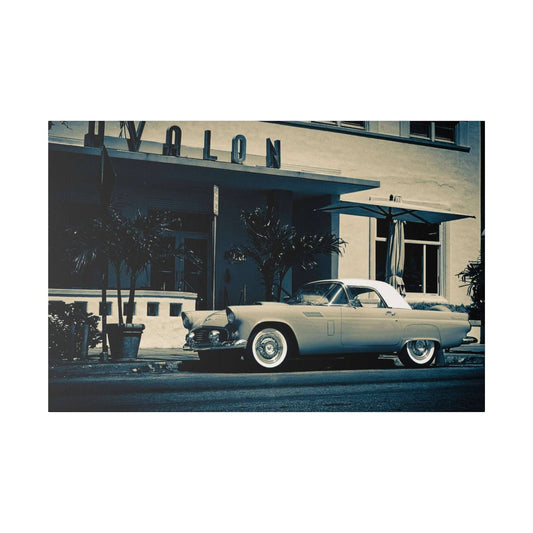 A vintage light-gray convertible with a white hardtop parked on a city street in front of a retro hotel with palm trees.