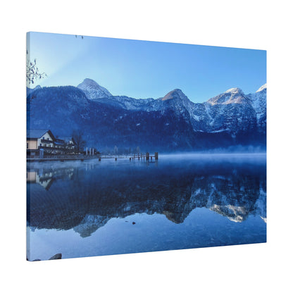 Snow-capped mountains rise above a calm lake with a wooden pier and a small village on the shore, reflecting in the water.