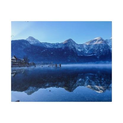 Snowy mountain range reflected in a calm blue lake, with a wooden pier and cottages on the left.
