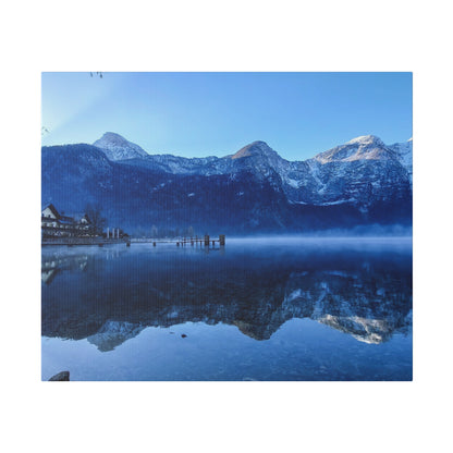 Snowy mountains rise above a calm lake with a small pier and houses on the left.