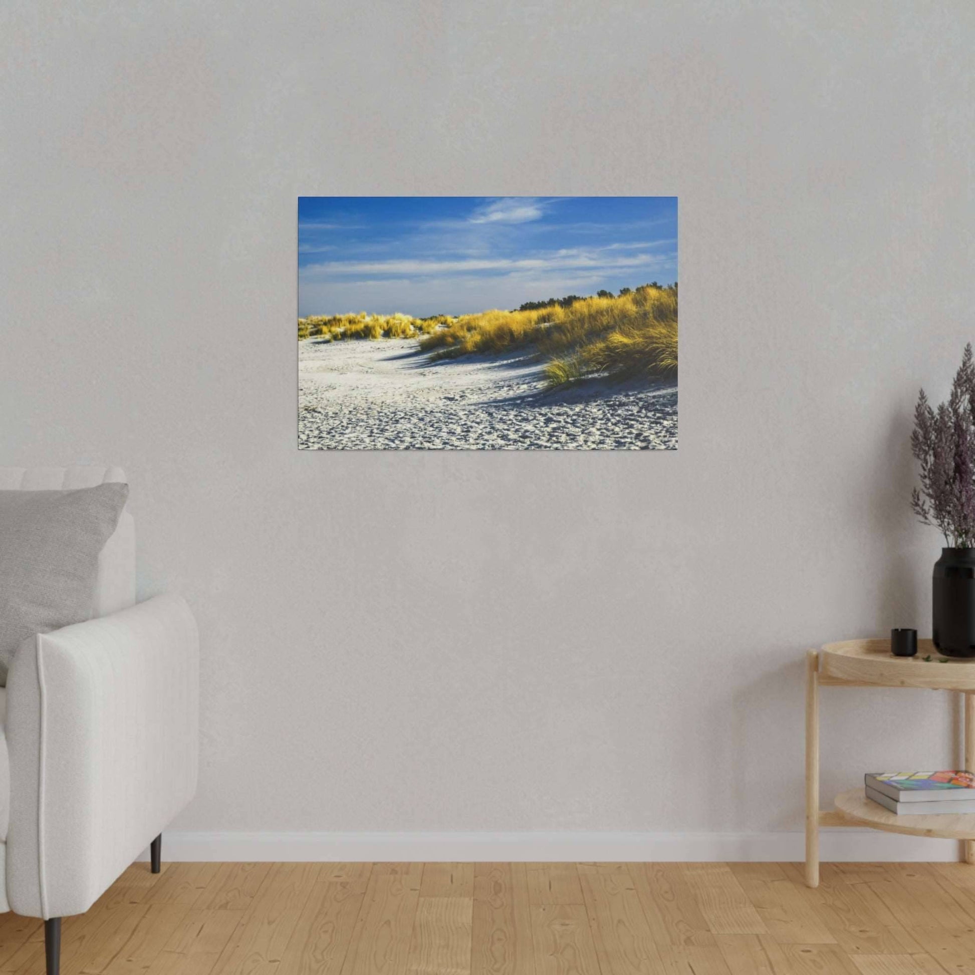 Framed photograph of sunlit sand dunes with yellow grasses under a blue sky, hung on a light gray wall above a wooden floor.