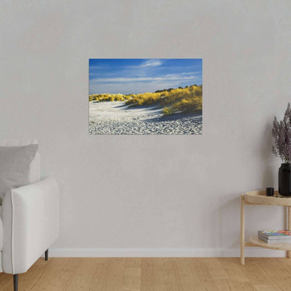 Framed photograph of sunlit sand dunes with yellow grasses under a blue sky, hung on a light gray wall above a wooden floor.