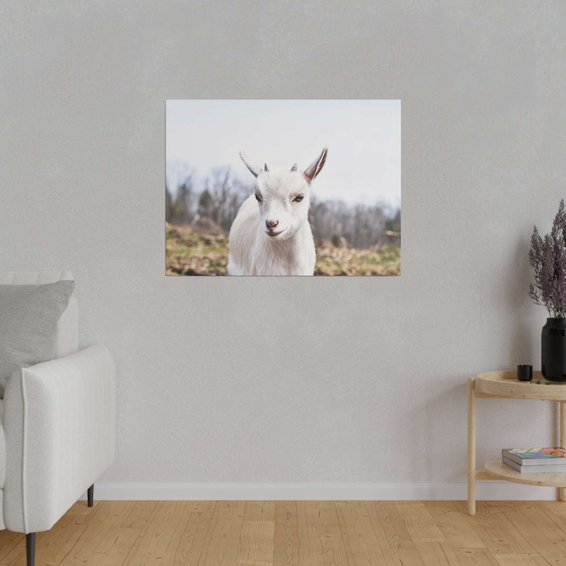 White goat portrait on a canvas, shown outdoors with a blurred field background, hung on a light gray wall.